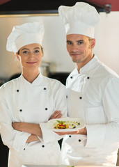 Two smiling chefs in a kitchen presenting a plate of fine food.