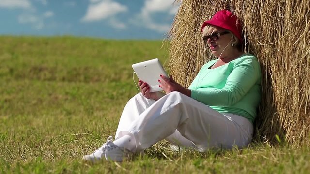 Senior blonde woman sits on a green grass near haystack and communicates via tablet computer. Female with tablet computer. Businesswoman with Tablet PC communicates through Skype