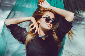Obraz premium young beautiful girl in sunglasses denim shorts and shirt posing while lying on a hammock, beautiful woman, brunette resting on vacation at the beach, relax, outdoor portrait