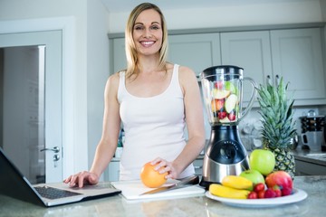 Obraz na płótnie Canvas Pretty blonde woman preparing a smoothie with recipe on laptop