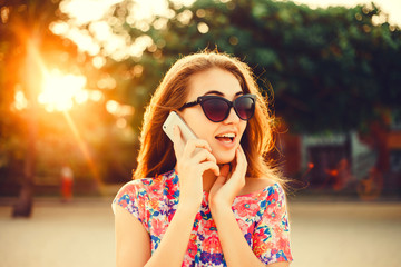 young beautiful brunette girl in sunglasses holding a smart phone, messages, posting photos to instagram, checks email,writes to friends SMS, outdoor portrait, close up, posing on the street at sunset