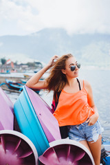 Fototapeta premium haired young girl in sunglasses and a red t-shirt denim shorts travels with a travel backpack for the world, posing next to a mountain lake, around the boats and boats