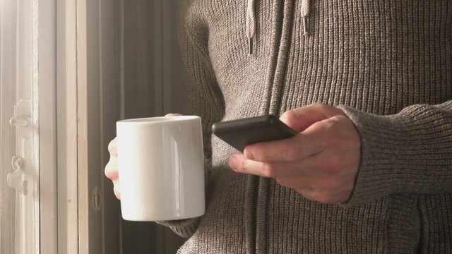Man drinking coffee and texting messages over smartphone, adult male standing by the window, using mobile smart phone for communication in the morning.