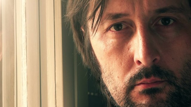Close up footage of sad adult male face, unshaven man standing by the window and thinking, looking at camera.