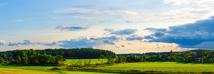 Rural landscape. Sunny summer evening. Sky with clouds, grass field, trees and farmhouses. Panoramic shot.