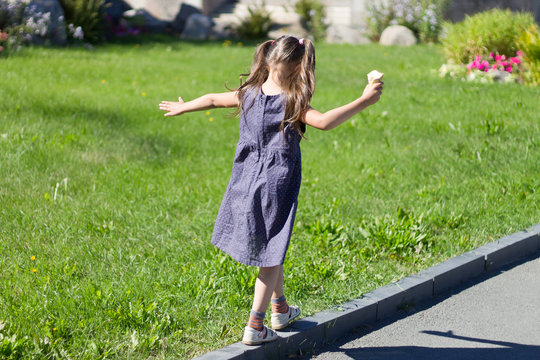 Little cute girl walking on the curb with ice cream in hand and intently trying to keep her balance, with her back