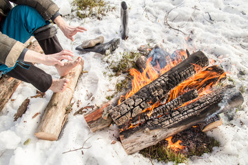 A man warms his hands and feet near the fire in the pine forest covered with snow in winter