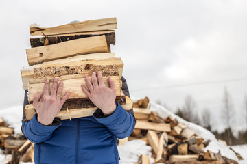 a man carries in his hand a great bunch of chopped firewood in the winter in the village against the backdrop of snow-covered heaps of other wood