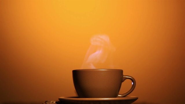 Brown cup with hot drink and coffee grains, steam, orange background.