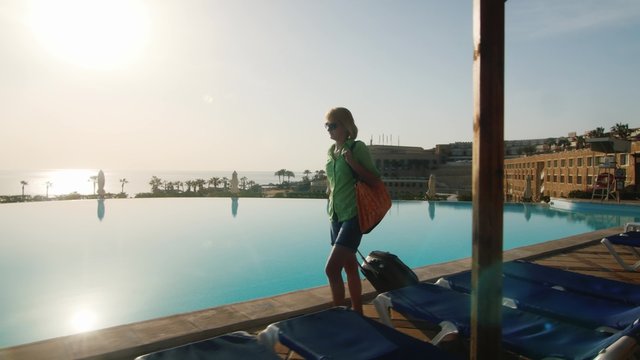 Steadicam shot of Woman with travel bag walking on recreation area along the pool