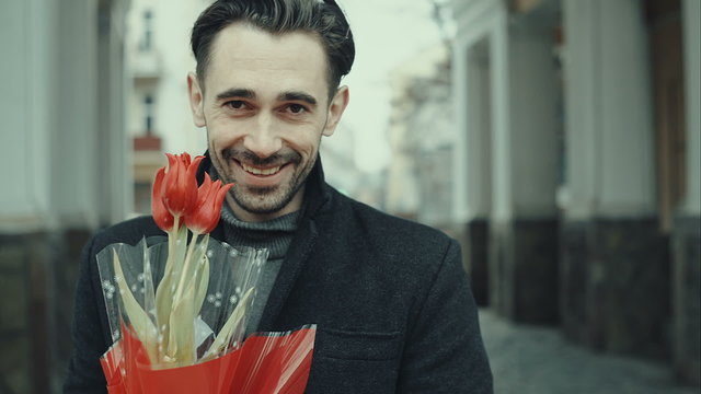 Happy smiling forty years old caucasian man with baguette and flower bouquet standing on the street. City buildings as background. RAW video rcord.