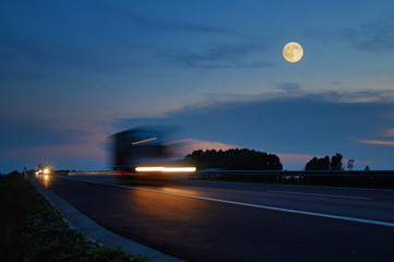 Trucks with glowing lights arriving on the motorway from afar in the landscape with a full moon at...