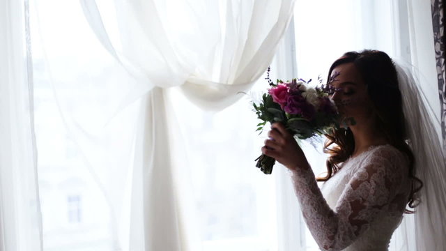 A bride of incredible beauty holding bridal bouquet at her left hand and smelling flowers aroma.