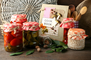 Jars with pickled vegetables and beans, spices, book of recipes and kitchen utensils on wooden background