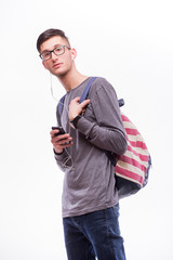 Happy hipster guy in glasses with backpack using a smart phone to listen music with headphones on white background