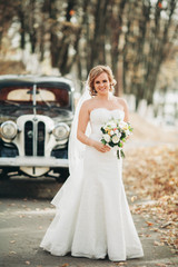 Fototapeta na wymiar Beautiful happy bride with bouquet near retro car in autumn
