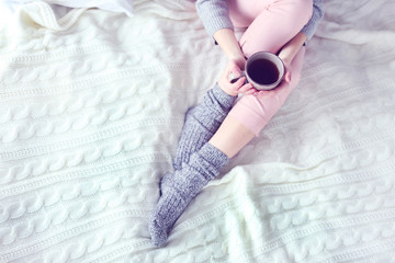 Woman in a pajamas and warm socks holding a cup of tea while sitting on a white knitted blanket