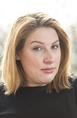 Classic Head Shot. A young woman in a black top and short blond hair stands outside back lit by the sun. She is looking in a slight questioning manner