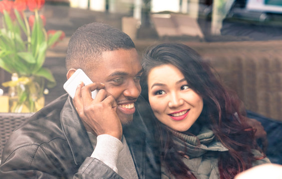 Multiracial couple on date view through cafe window - Afro american man using phone and asian girlfriend facial expression in love - Cheerful mixed race friends - Closeup from glass focus on male 