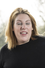 Classic Head Shot. A young woman in a black top and short blond hair stands outside back lit by the sun. She had been shaking her head and is not standing quiet with her eyes closed.