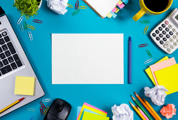 Office table desk with set of colorful supplies, white blank note pad, cup, pen, pc, crumpled paper, flower on blue background. Top view and copy space for text