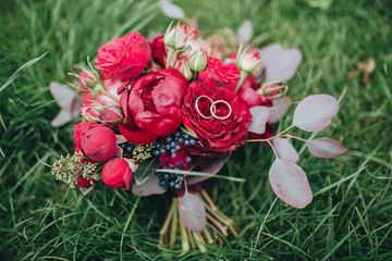 The bride's bouquet. Wedding bouquet . Wedding rings. A bouquet of red and pink flowers with black berries and greens with a ribbon Marsala color lies on a green lawn. Wedding rings lie on a bouquet