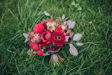 Obraz na płótnie Canvas The bride's bouquet. Wedding bouquet . Wedding rings. A bouquet of red and pink flowers with black berries and greens with a ribbon Marsala color lies on a green lawn. Wedding rings lie on a bouquet