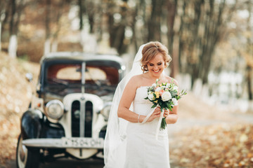 Beautiful happy bride with bouquet near retro car in autumn