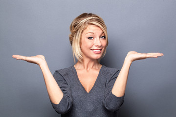 comparison concept - happy young blond woman displaying something on both flat hands for similar choice of product, gray background studio. - 105630602