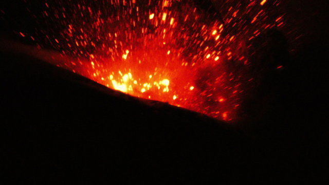 A powerful nighttime eruption of the Tungurahua volcano in Ecuador, bright red and orange lava and molten rock exploding from the crater and cascading down the dark slopes against a black sky.
