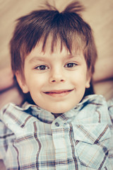 Closeup portrait of smiling little boy with brown eyes wearing checkered shirt lying on floor and looking at camera. Happy childhood concept, selective focus on eyes, top view, instagram filters