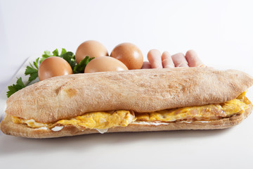 Sandwich in a bun with scrambled eggs with ham and cheese over the sour cream, set next to fresh eggs and ham.