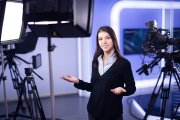 Television presenter recording in television news studio.Female journalist anchor presenting business report.News camera,light equipment behind the scenes.Talking at camera to the TV audience - obrazy, fototapety, plakaty