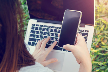 Closeup image of a woman hand holding smartphone with laptop background on green grass. at park outdoor and sunlight.