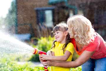 Little gardener girl with mother watering on lawn near house