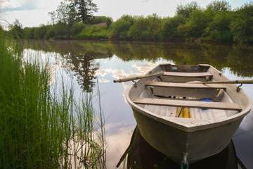 Rowboat in a narrow river. Blue sky and white clouds reflected in the river. There are bushes and trees on river banks.