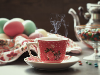 A cup of hot tea. Above the vapor from cup of hot beverage. In the background, easter eggs, kettle