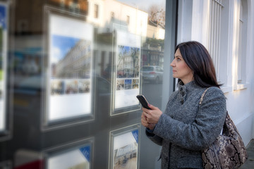 Mature woman wearing a grey coat and a purse is looking at the ads in the window of a real estate...
