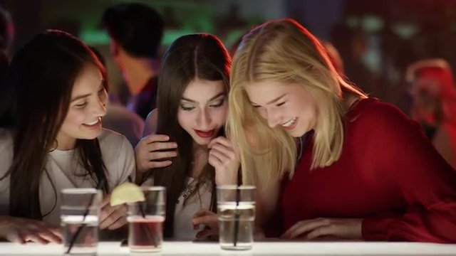 three girls at bar in club looking at smartphone and laughing