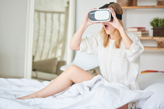 The girl in white sitting on the bed in the virtual reality helmet