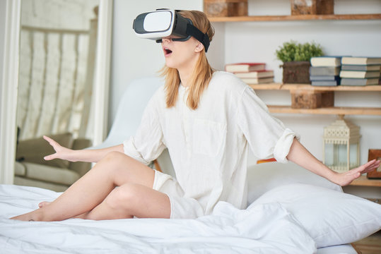 The girl in white sitting on the bed in the virtual reality helmet