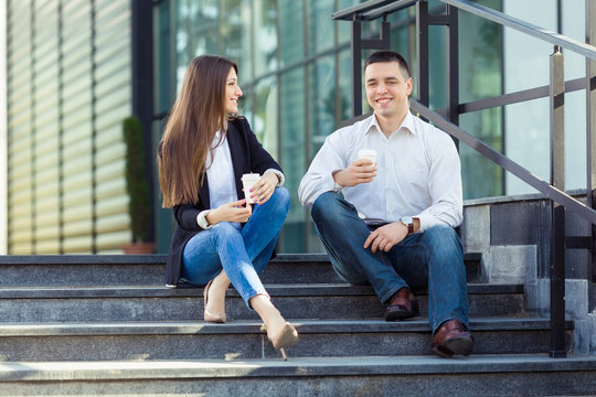 Young businesswoman and businessman sitting on the steps of the office building, drinking coffee and talking. Young man is looking at camera and young woman is looking at him.