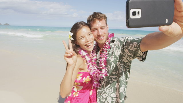 Couple on beach taking selfie with smartphone.  Young woman and man in love using mobile cell smart phone on Hawaii beach vacation in Hawaiian clothing wearing Aloha shirt dress and flower lei.