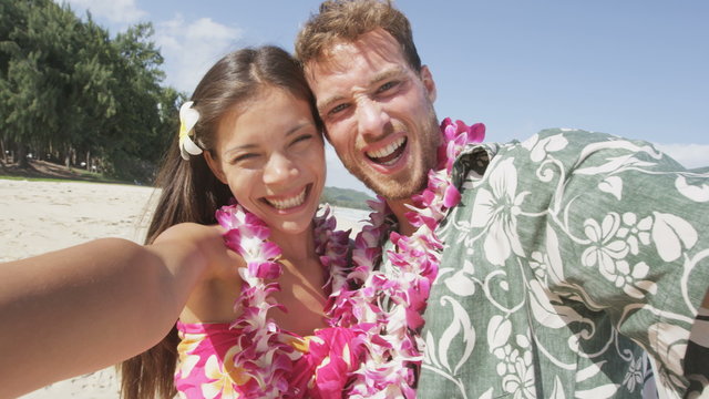 Couple on beach taking selfie photo on Hawaii with smart phone.  Young woman and man in love using smartphone on beach vacations in Hawaiian clothing wearing Aloha shirt dress and flower lei. RED EPIC