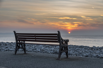 An empty bench, at the see shore, with a fiery, colourful sun setting into the ocean