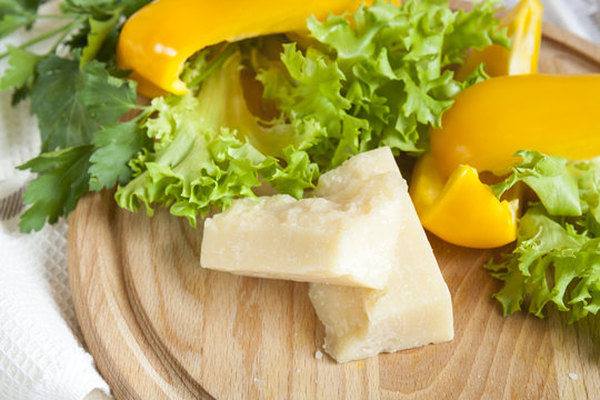 Parmigiano (parmesan) cheese around fresh yellow pepper lettuce leaves parsley on wooden board and napkin background