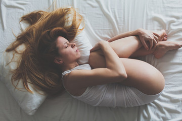 Young woman lying down on the bed in fetus position - obrazy, fototapety, plakaty