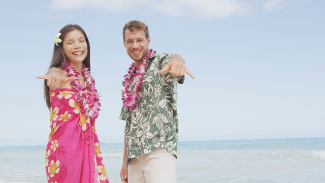 Portrait of couple of tourists happy standing on Hawaiian beach at their Hawaii vacation. Asian woman and Caucasian man wearing flower lei garland and Aloha clothing showing Shaka hand sign on travel.