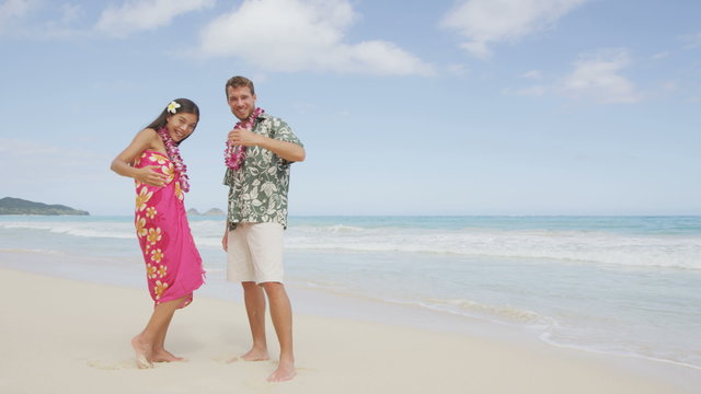 Hawaii beach couple saying welcome and come here showing hand gesture waving hand gesturing. Portrait of Asian woman and Caucasian man on beach Aloha Hawaiian shirt with flower leis and typical attire