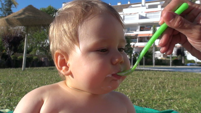 blonde baby one year old eating yogurt from green plastic spoon woman hand in green grass garden next to swimming pool on summer holidays